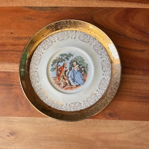 May include: Vintage decorative plate with a gold rim and a central image of a couple in period clothing. The plate has a white background with gold filigree detailing. The outer rim is a wide band of gold.