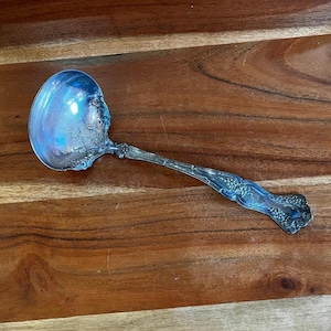 May include: A silver ladle with ornate floral and vine detailing. The ladle's bowl is round and reflective, and the handle features a decorative pattern. The utensil is set against a wooden surface.