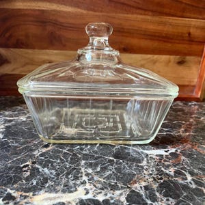 May include: A clear glass butter dish with a rectangular base and a matching lid. The lid features a small knob handle at the top, and both the base and lid have vertical ribbed detailing along the sides. The base has a geometric pattern.