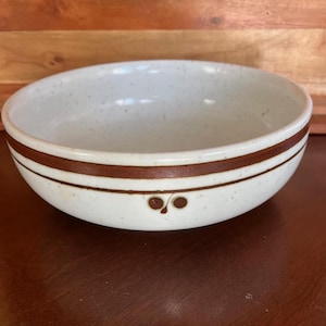 May include: A round ceramic bowl with a speckled off-white finish. The exterior features two horizontal brown bands encircling the middle, with a small, stylized brown leaf or berry motif centered below the lower band on the side of the bowl.