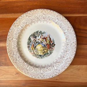 May include: A vintage decorative plate with a cream-colored base and gold filigree border. The center features a colorful illustration of a couple in period clothing dancing, with two other figures nearby. The plate is likely made of ceramic.