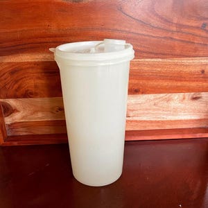 May include: A tall, off-white plastic tumbler with a matching lid and a small spout. The tumbler is cylindrical and appears to be empty. The lid has a small opening for drinking. The background is a wooden surface.