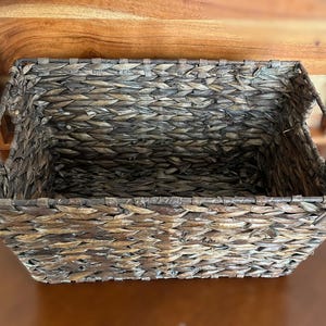 May include: A rectangular storage basket woven from dark brown natural fibers. The basket features a sturdy, open-top design with integrated side handles, showcasing a detailed, multi-directional braided pattern throughout the entire structure.