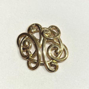 May include: A gold-toned brooch with an intricate, swirling design. The brooch features a series of interconnected, looping shapes, creating a decorative and elegant accessory. The pin is a classic piece of jewelry.