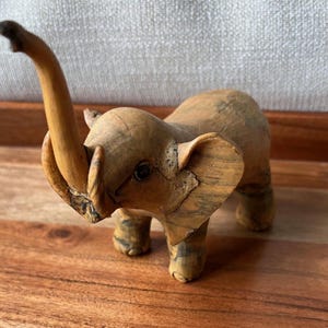 May include: A wooden elephant figurine with a raised trunk and large ears. The elephant is light brown with darker brown markings and is standing on four legs. The figurine is approximately 4 inches tall.