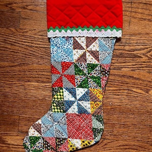 May include: A Christmas stocking featuring a quilted red cuff with a green scalloped trim and white eyelet lace. The body of the stocking is constructed from a patchwork of various colorful fabric squares, each containing small geometric patterns.