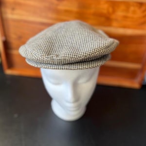 May include: A flat cap featuring a small-scale houndstooth pattern in shades of beige, brown, and white. The cap has a rounded crown and a short, stiff brim that extends from the front, displaying a structured shape and a woven fabric texture.