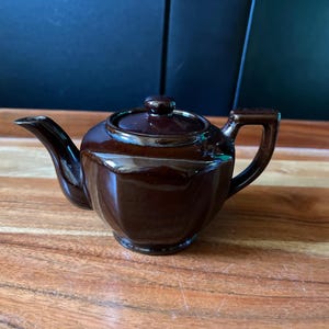 May include: A small, dark brown ceramic teapot with a glossy finish. The teapot features a hexagonal body shape, a curved spout, a matching lid with a small knob handle, and a sturdy, angular handle on the opposite side of the spout.