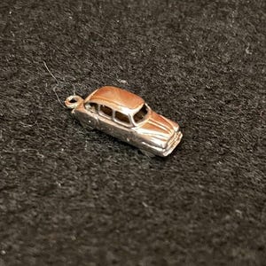May include: A small, silver-colored metal charm shaped like a vintage sedan car. The charm features a detailed roofline, window cutouts, and a hood design, with a small circular loop attached to the front for hanging or attachment to a chain.