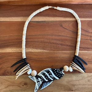 May include: A necklace featuring a central black and white fish pendant, flanked by wooden beads and clusters of black and tan pointed spikes. The strand consists of small, circular, off-white disc beads, creating a layered and textured appearance.