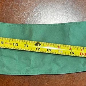 May include: A green fabric item, possibly a scarf or accessory, is measured with a yellow tape measure. The tape measure shows a length of approximately 25 inches. The fabric is a solid, medium green color.