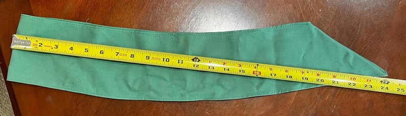 May include: A green fabric item, possibly a scarf or accessory, is measured with a yellow tape measure. The tape measure shows a length of approximately 25 inches. The fabric is a solid, medium green color.