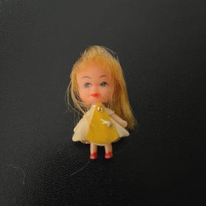 May include: A small doll with blonde hair, blue eyes, and red shoes. The doll wears a yellow dress with a white collar and sleeves. The doll is a miniature collectible.