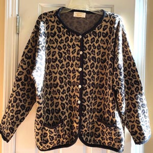 May include: A button-down cardigan sweater with a leopard print pattern in shades of tan, brown, and black. The sweater has a navy blue trim, two front pockets, and white buttons. The garment is hanging on a white hanger.