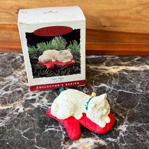 May include: A white cat figurine resting on a red quilted mitten clip-on ornament. The packaging box features the Hallmark Keepsake logo, the text Cat Naps CLIP-ON, and the text COLLECTOR'S SERIES. The cat has a green collar with a gold bell.