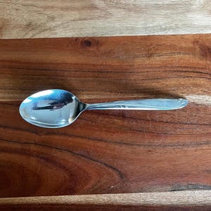 May include: A shiny, silver spoon with a rounded bowl and a long, slender handle. The spoon is resting on a wooden surface with a rich, brown grain pattern. The spoon reflects the surrounding environment.