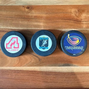 May include: Three black hockey pucks with team logos. One puck has a red and white flame design with the word "Flames". Another has a hockey player silhouette. The third has a blue and purple design with the word "Thrashers".