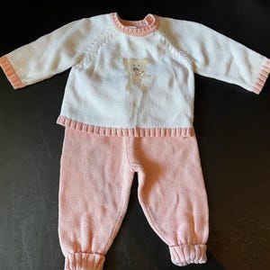May include: A two-piece baby outfit in white and pink. The long-sleeved top has a teddy bear design on the front. The outfit is made of a soft knit material, with pink cuffs and trim. The pants are pink with ribbed cuffs.