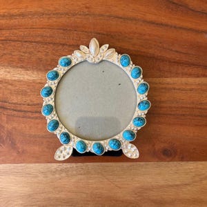 May include: A round silver-toned picture frame featuring a border of oval turquoise-colored stones and small white accents. The frame has a decorative top crest and two bottom feet, surrounding a circular gray center area for displaying a photograph.
