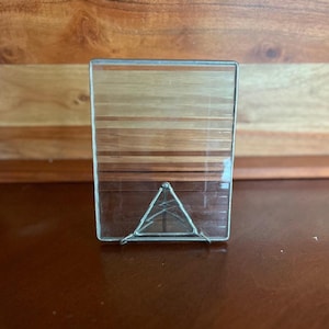 May include: A clear glass box with a wooden panel design on the top half. The bottom half features a triangular metal design. The box is rectangular and has a silver metal frame.