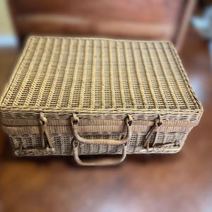 May include: A rectangular, light brown wicker picnic basket with a woven pattern. The basket has a handle and two metal clasps for closure. The basket is sitting on a brown surface.