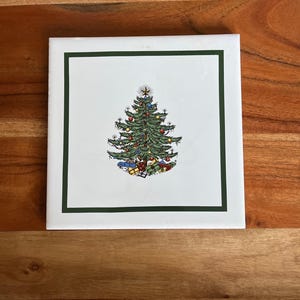 May include: Square ceramic tile featuring a detailed illustration of a decorated Christmas tree. The tree is green with colorful ornaments and topped with a star. Presents are at the base of the tree. The image is framed by a green border.