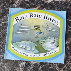 May include: A children's picture book titled Rain Rain Rivers with words and pictures by Uri Shulevitz. The cover features an illustration of a person running across a landscape with puddles, birds, and clouds, framed by a light blue and lime green border.