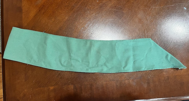 May include: A seafoam green fabric collar, likely for a garment, lies on a brown wooden surface. The collar is rectangular with a pointed end, and appears to be made of a soft, woven material. The item is a solid color.