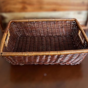 May include: A rectangular brown wicker basket with handles on each end. The basket is made of woven brown wicker and has a rectangular shape. The basket is empty and sits on a brown surface.