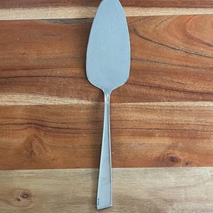 May include: A stainless steel cake server with a serrated edge and a long, slender handle. The server is designed for cutting and serving cake. The server is shown on a wooden surface.