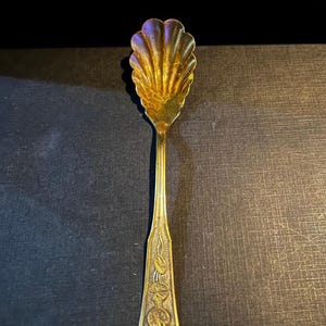 May include: A vintage gold-colored spoon with a shell-shaped bowl and a long, decorated handle. The spoon's bowl has a textured, fan-like design. The handle features floral and geometric patterns, adding to its antique charm.