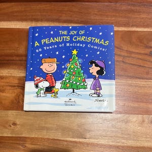 May include: A hardcover book titled "The Joy of a Peanuts Christmas" featuring Snoopy, Charlie Brown, and Lucy. The cover shows a decorated Christmas tree and a snowy background. The book celebrates 50 years of Holiday Comics.