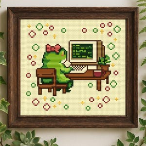 May include: Cross-stitch art of a green frog with a pink bow working on a computer. The frog is seated at a brown desk with a monitor, keyboard, and a small plant. The background is a light beige with decorative green and red shapes. The artwork is framed in a dark brown wooden frame.