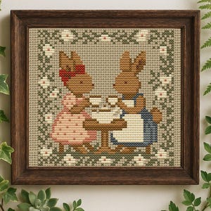 May include: Cross-stitch framed artwork featuring two brown bunnies at a table, drinking tea. One bunny wears a pink dress and red bow, the other a blue apron. The background is a light green with floral accents, all within a dark brown wooden frame.