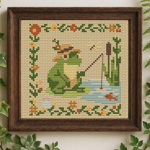 May include: Cross-stitch art of a green frog fishing in a pond, wearing a hat. The artwork is framed in a dark brown wooden frame. The scene includes a fish, cattails, and a dragonfly. Floral border.