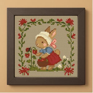 May include: Cross-stitch framed artwork featuring a bunny picking strawberries. The bunny wears a white bonnet, blue shirt, and red skirt. The frame is dark brown, and the background is a neutral beige. A floral border surrounds the bunny.
