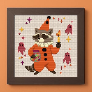 Op de afbeelding: Kruissteekkunstwerk van een wasbeer in een oranje clownspak, met een kaars en een pot kleurrijke snoepjes. Het ingelijste stuk heeft een grillig ontwerp met ster- en spookmotieven, perfect voor Halloween-decoratie.