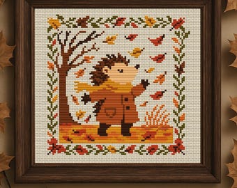 May include: Cross-stitch artwork in a wooden frame depicts an autumn scene. A hedgehog in a coat and scarf stands in a field of fallen leaves, with a tree and falling leaves in the background. The border features leaves and vines.