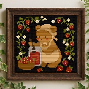 Op de afbeelding: Kruissteekkunst van een beer die jam maakt, omringd door aardbeien en bloemaccenten. De beer draagt een muts en een schort, met een pot jam en een mand met aardbeien. Het kunstwerk is ingelijst in een donkerbruine houten lijst.