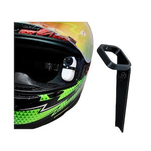 Peut inclure: Casque de moto noir et vert avec une caméra blanche montée et un support noir séparé. Le casque a une visière et un motif géométrique vert et noir. La caméra est fixée sur le côté du casque.