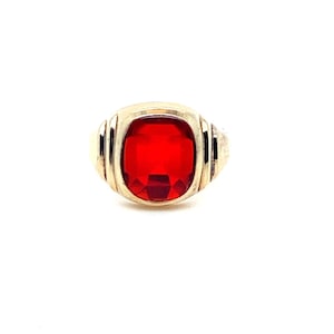 May include: A gold-toned ring featuring a large, rectangular, faceted red gemstone set in the center. The band displays a series of vertical grooves or ridges on both sides of the central stone, creating a structured and symmetrical appearance.