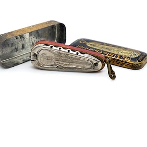 May include: Vintage harmonica with a red and silver body, featuring a violin-shaped design. The harmonica is next to its rectangular tin case, which has a gold and black label that reads "My Little Charmer". The case is open, revealing the harmonica.