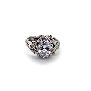 May include: A silver ring with an oval, clear gemstone. The ring has an ornate, filigree design around the gemstone. The ring is set against a white background.