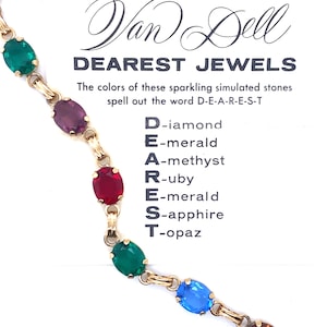 May include: A gold-toned necklace with oval-shaped simulated gemstones in emerald green, amethyst purple, ruby red, sapphire blue, and topaz. The text reads "Van Dell DEAREST JEWELS". The colors spell out the word D-E-A-R-E-S-T.