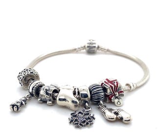May include: A silver charm bracelet with various holiday-themed charms. Charms include a snowflake, a stocking, a gift box, and a pair of mittens. The bracelet is made of a flexible, snake-like chain.