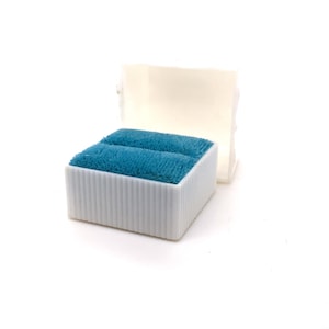 May include: A white, rectangular ring box with a ribbed exterior and a teal velvet interior. The box is open, revealing two slots for rings. The lid is propped up behind the box, against a white background.