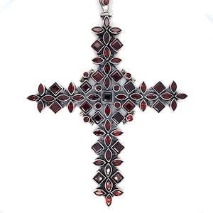 May include: A large, ornate silver-toned cross pendant with numerous deep red gemstones. The cross features a central square stone, surrounded by various shapes of red gems, including squares, ovals, and round stones, set in a detailed, floral-like pattern.
