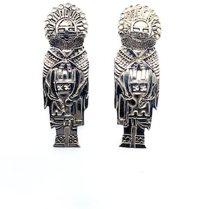 May include: A pair of silver-toned earrings. Each earring features a detailed design of a figure with a headdress, intricate facial features, and a patterned body. The earrings are displayed against a white background.