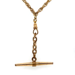 May include: A gold-toned watch chain necklace featuring a central T-bar pendant suspended from a short vertical chain link section. The main chain consists of twisted links that meet at a small connector, leading down to the T-bar drop component.
