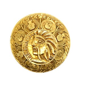 May include: A gold-toned, circular brooch with a central relief of a face and surrounding coin-like details. The brooch has a detailed, textured design and is set against a white background.